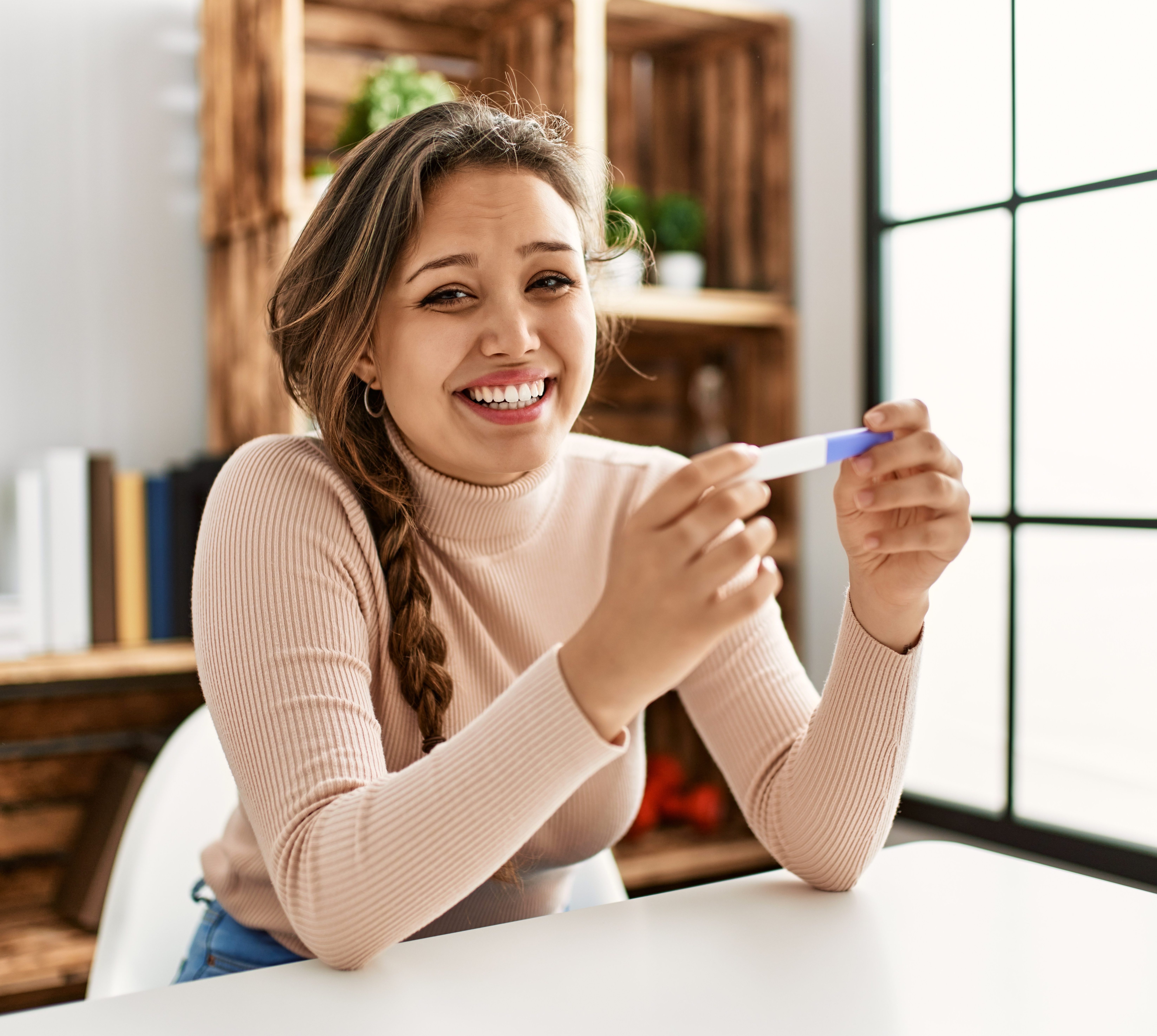 Young beautiful hispanic woman smiling confident holding pregnancy test at home